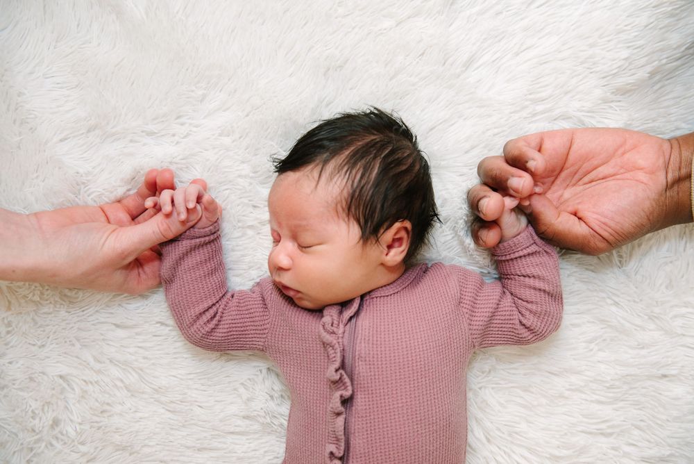 A sleeping baby in a pink ribbed onesie holds hands with adults while lying on a white fluffy blanket.