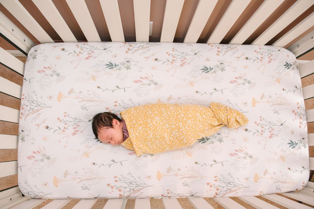 A sleeping infant wrapped in a yellow blanket lies on a floral patterned mattress in a wooden crib.