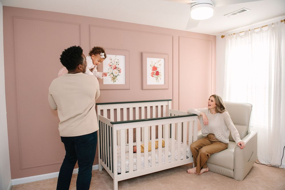 Modern nursery room with blush pink walls, white furniture, and natural light streaming through sheer curtains.
