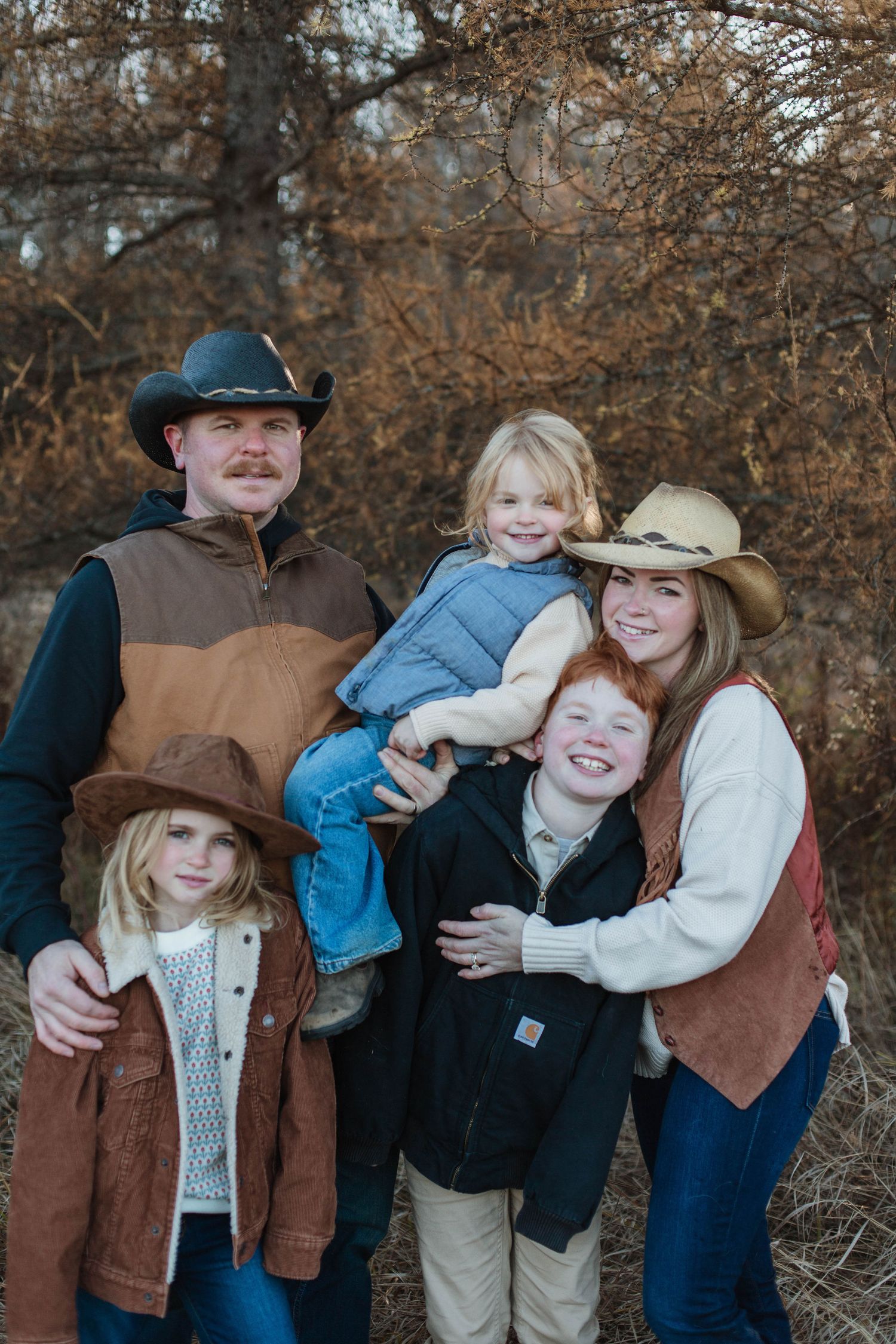 A group in western wear poses outdoors for a casual family portrait during autumn with brown foliage in background.