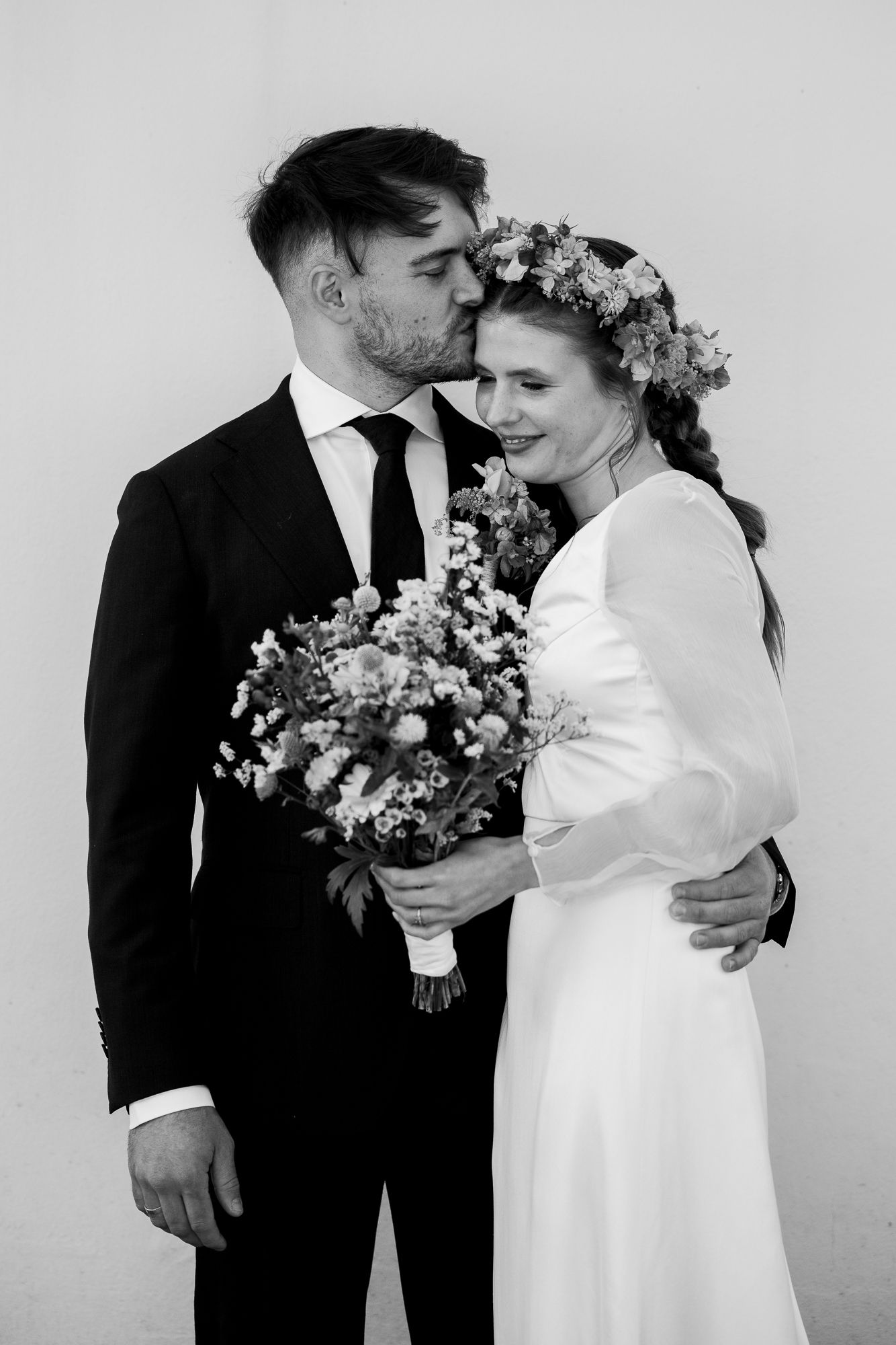 Black and white wedding photo of bride in floral crown and minimal dress with groom in black suit holding bouquet.