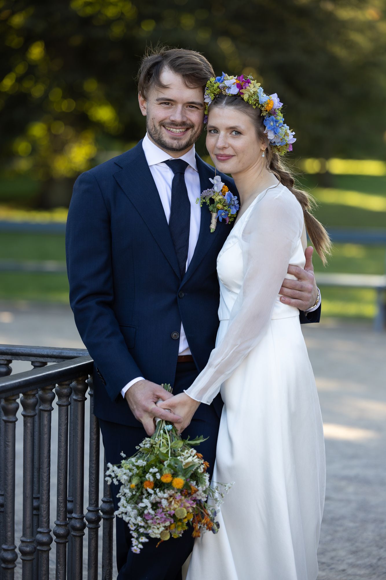 A wedding couple stands together outdoors with the bride wearing a flower crown and holding a rustic wildflower bouquet.