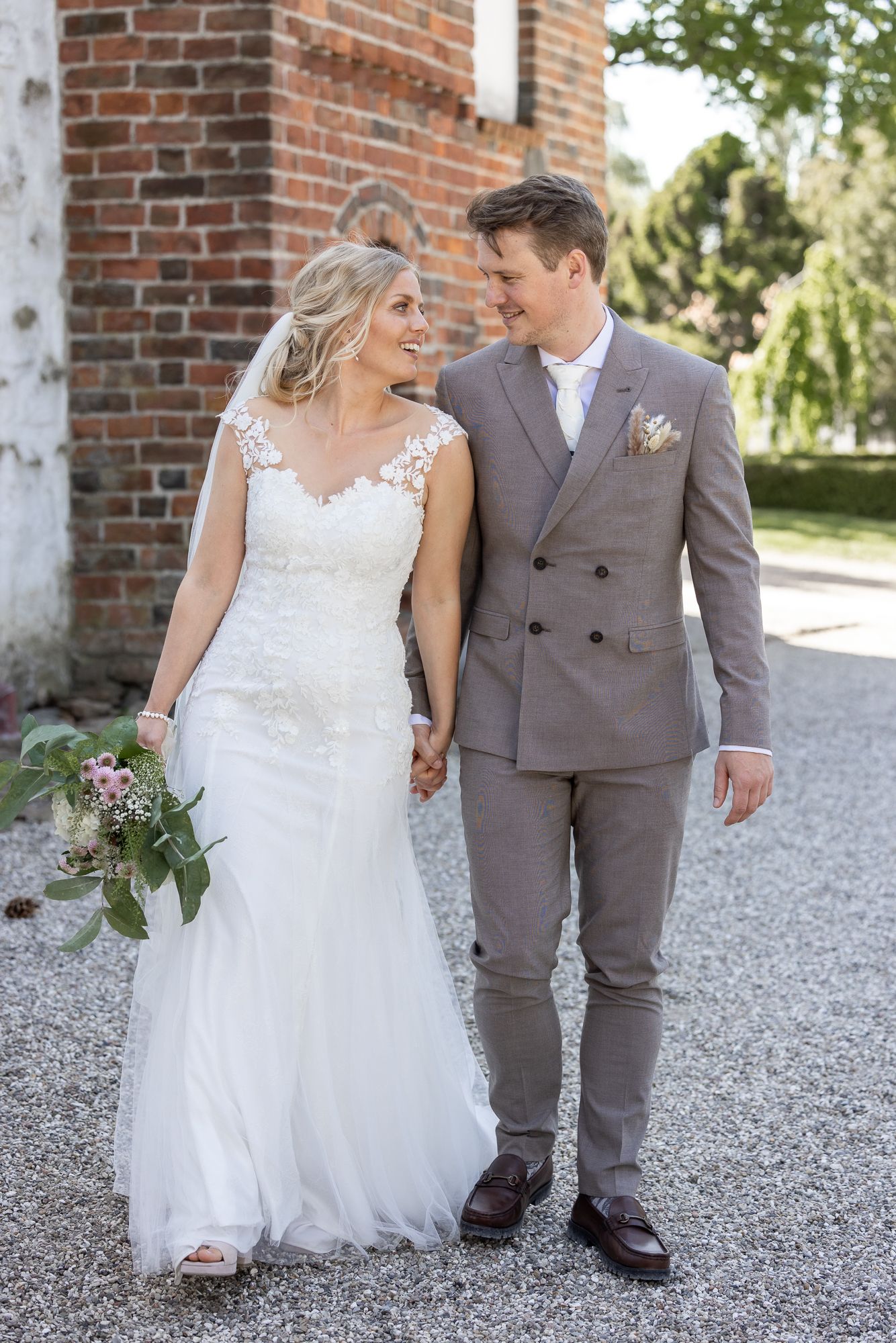 Newlyweds share a loving moment while walking on a gravel path near a rustic brick building.