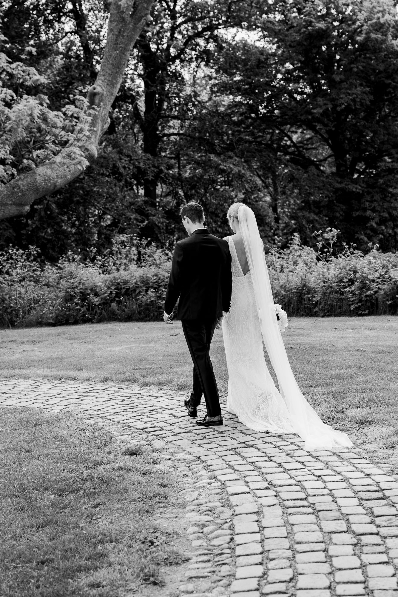 A couple walks down a winding stone path surrounded by lush garden foliage in black and white.