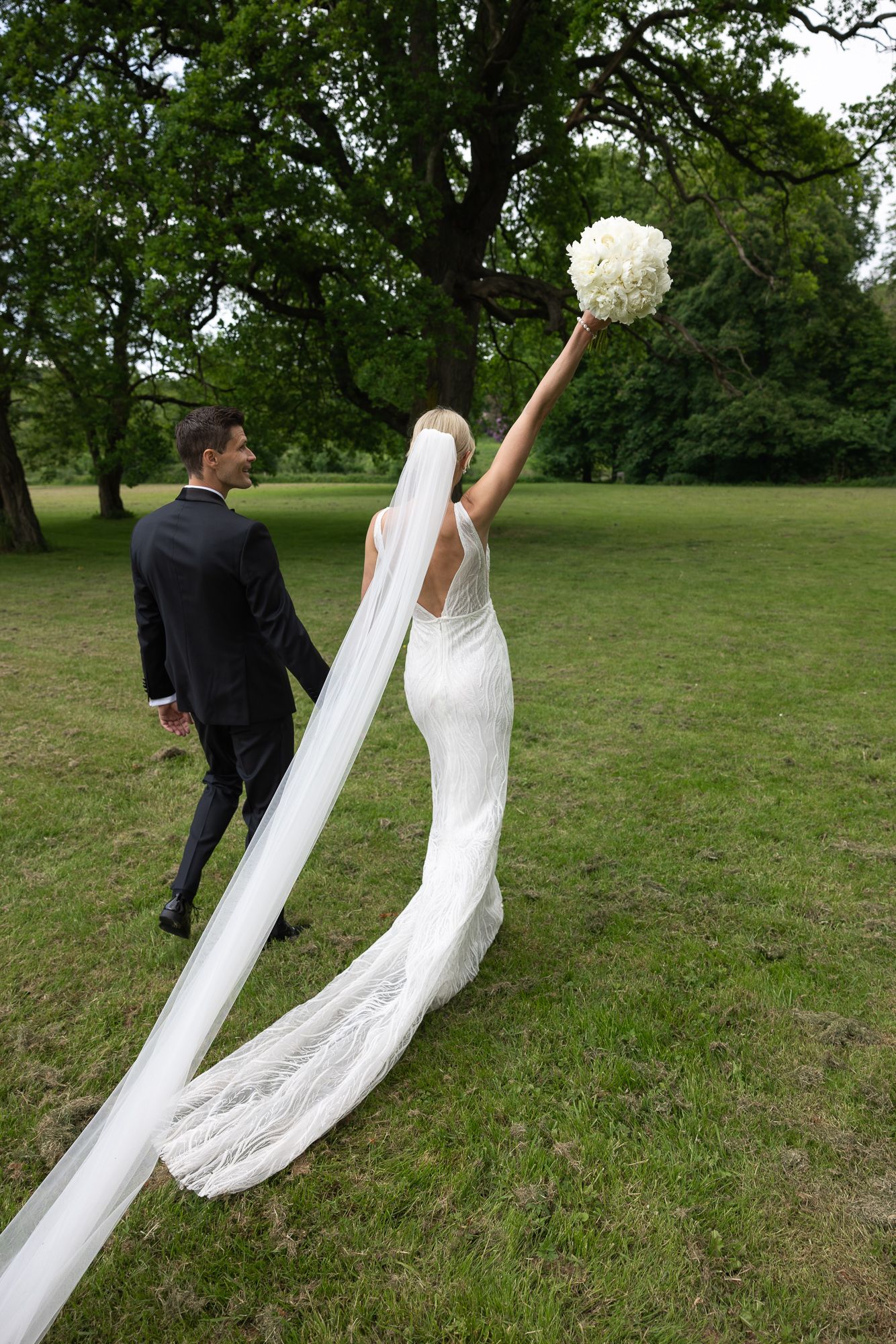 A couple in wedding attire poses playfully in a garden setting with a raised white bouquet.