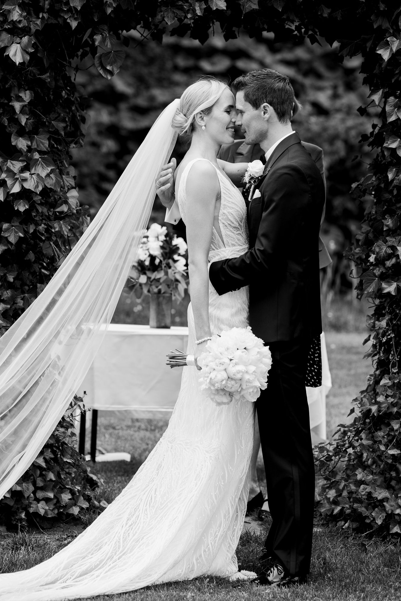 A romantic wedding portrait captured under an ivy-covered archway in black and white.