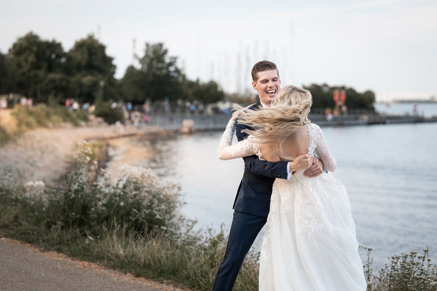 Playful wedding couple embracing by waterfront at sunset.