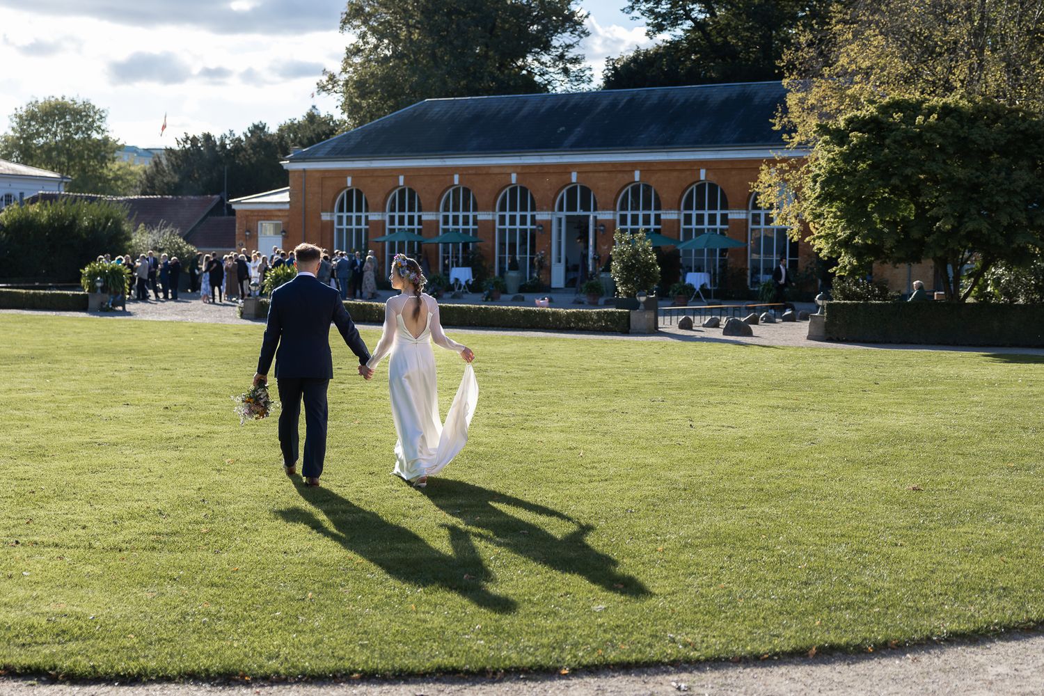 A wedding couple walks hand in hand across a sunny lawn towards Brøndsalen Frederiksberg Have.