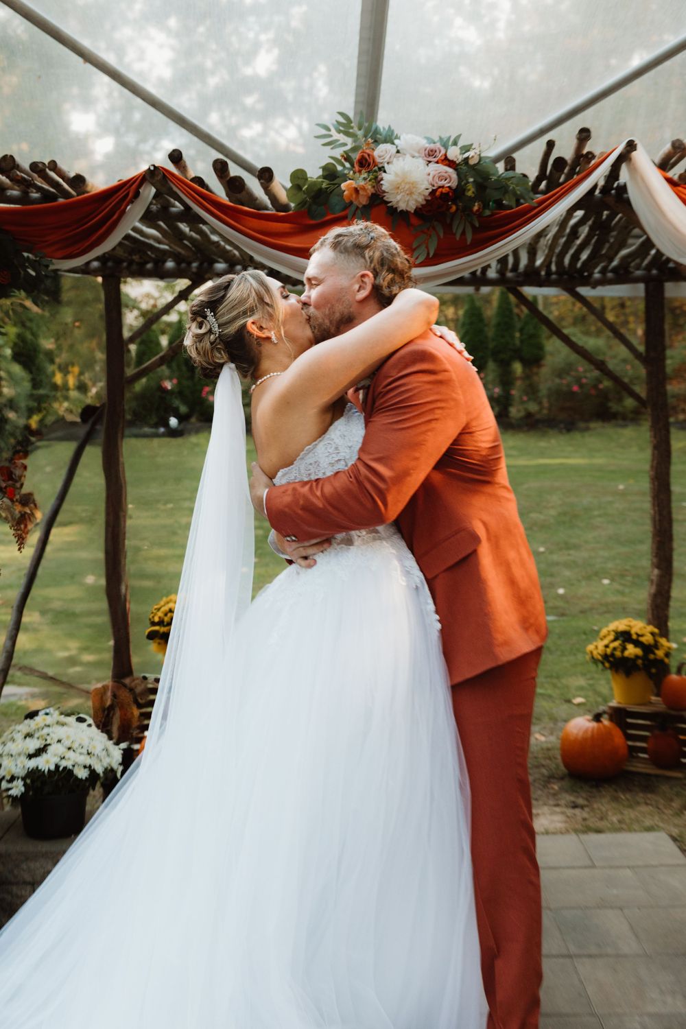 Wedding couple embracing under a rustic wooden pergola decorated with autumn flowers.
