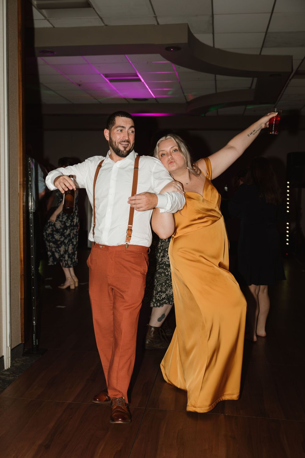 Wedding party dancing energetically on wooden dance floor with colorful lighting.