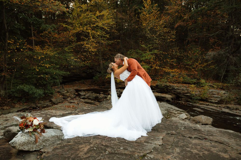 Dramatic wedding photo on rocky terrain with flowing white dress against forest backdrop.