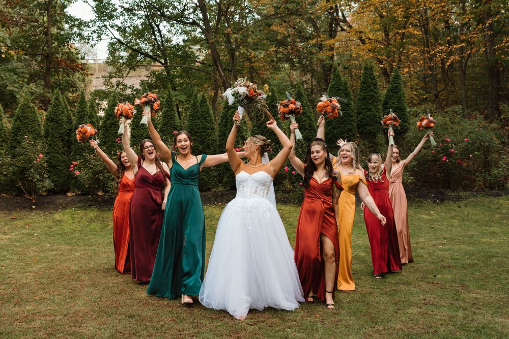 Wedding party in white, burgundy and emerald dresses poses on autumn lawn.