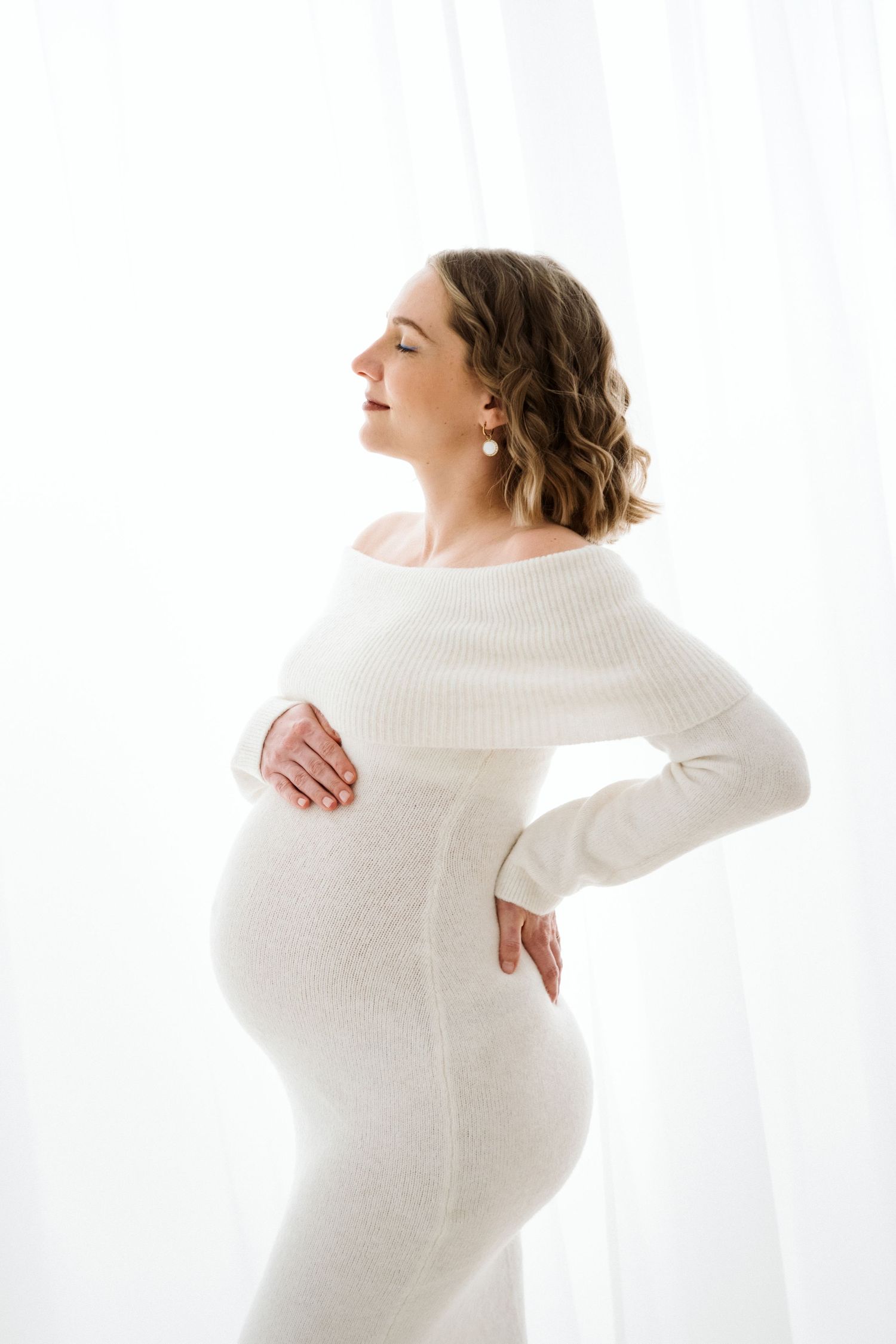 Artistic maternity photo series in white showing a pregnant woman in a sweater against a bright background.