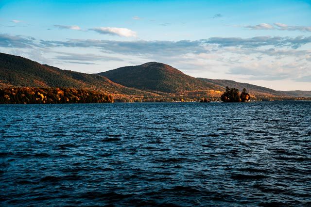 Mountain lake with rippling dark waters surrounded by autumn-colored hills under a blue cloudy sky.