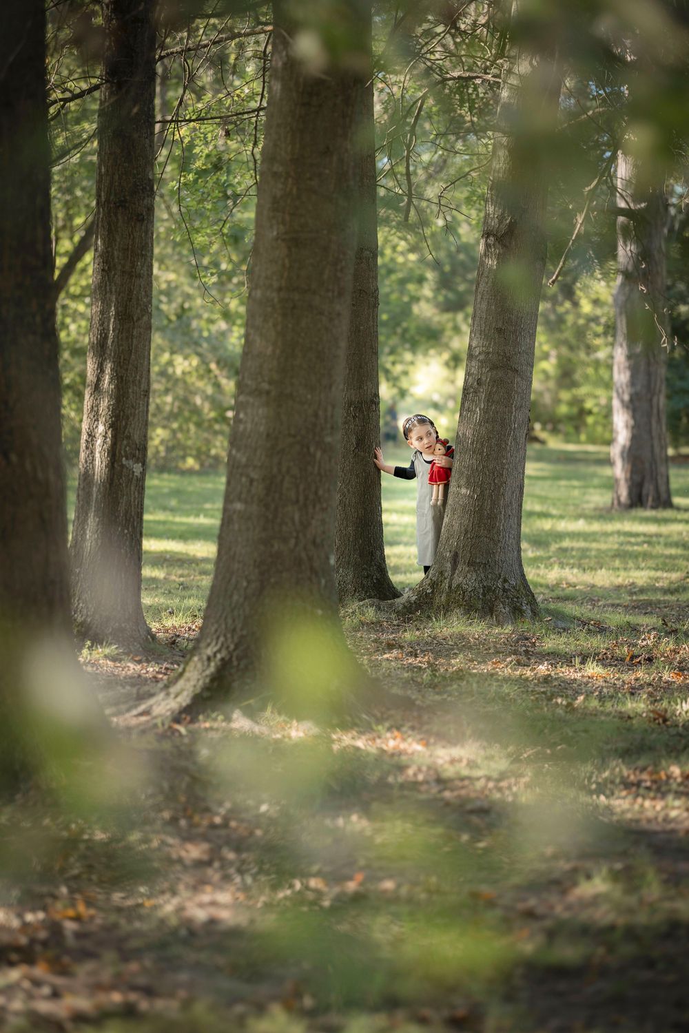 little girl peeking playfully from behind a large tree trunk in a serene wooded park area on a sunny day.