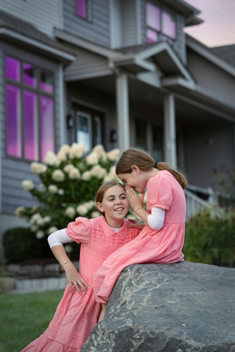 Two people in matching coral pink dresses sit together on a large rock at dusk with a house in the background.