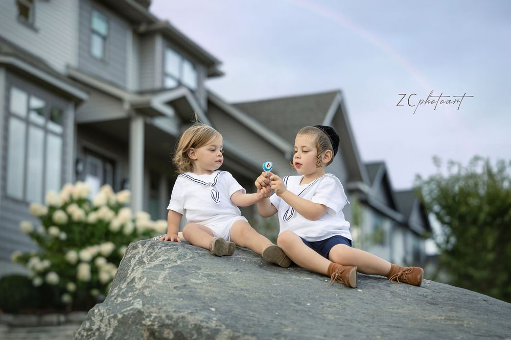 Two young children in white shirts sit on a large rock having an animated conversation outside a suburban house.