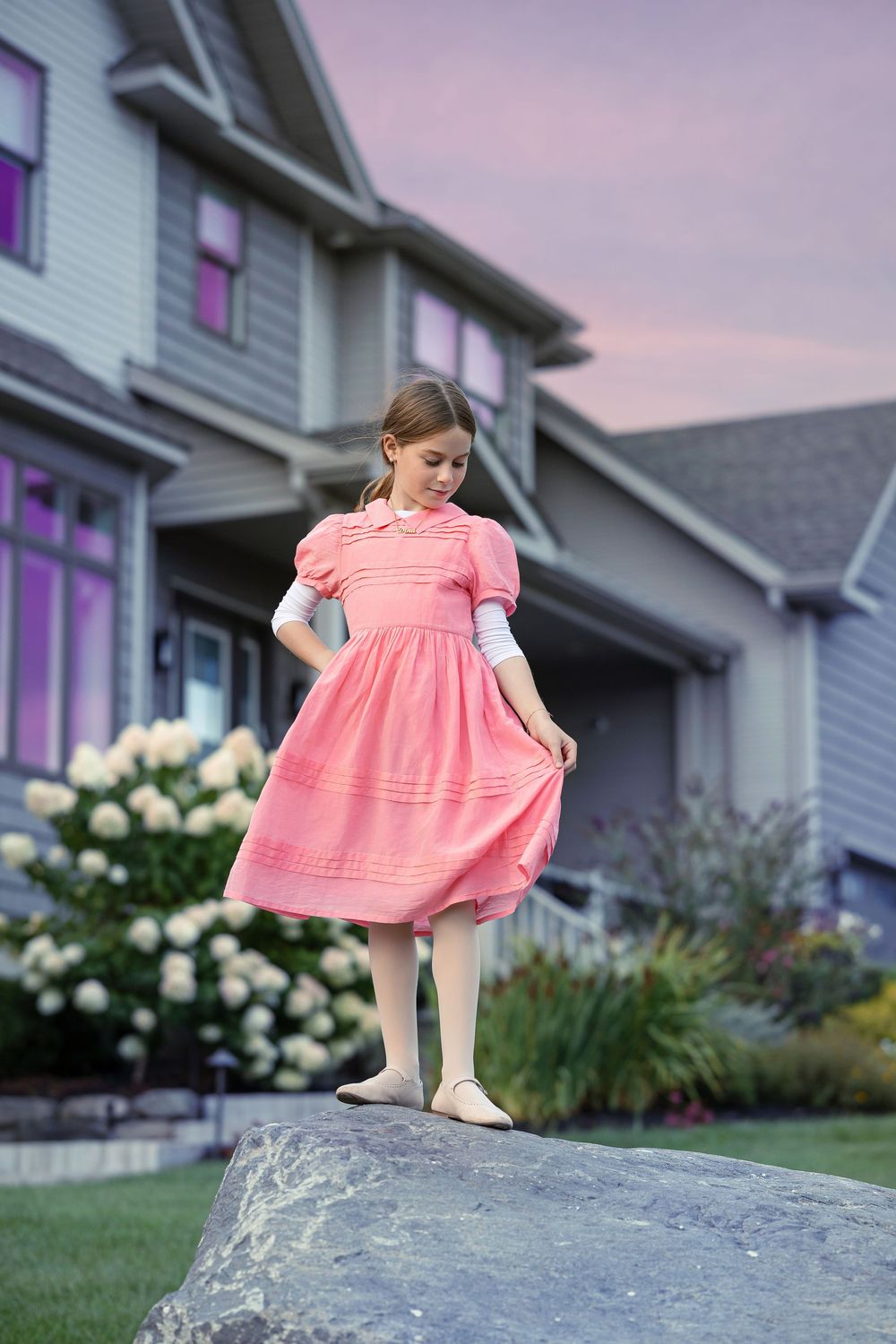 A young child in a pink dress plays and climbs on a large rock formation in front of a suburban home at dusk.