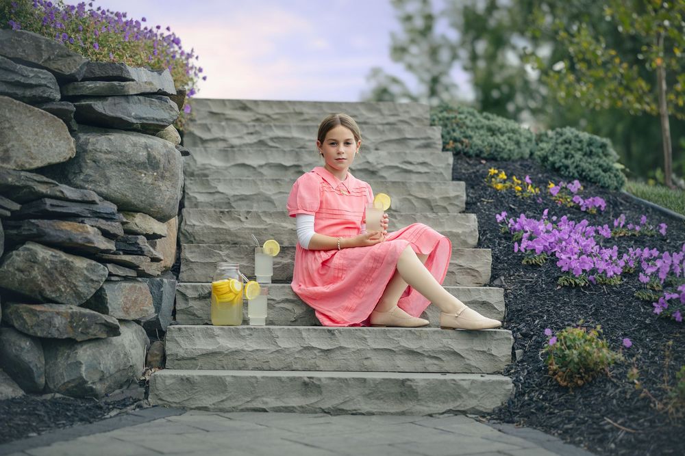 A pink-clad figure sits on a rustic stone wall surrounded by blooming purple flowers in a garden setting.