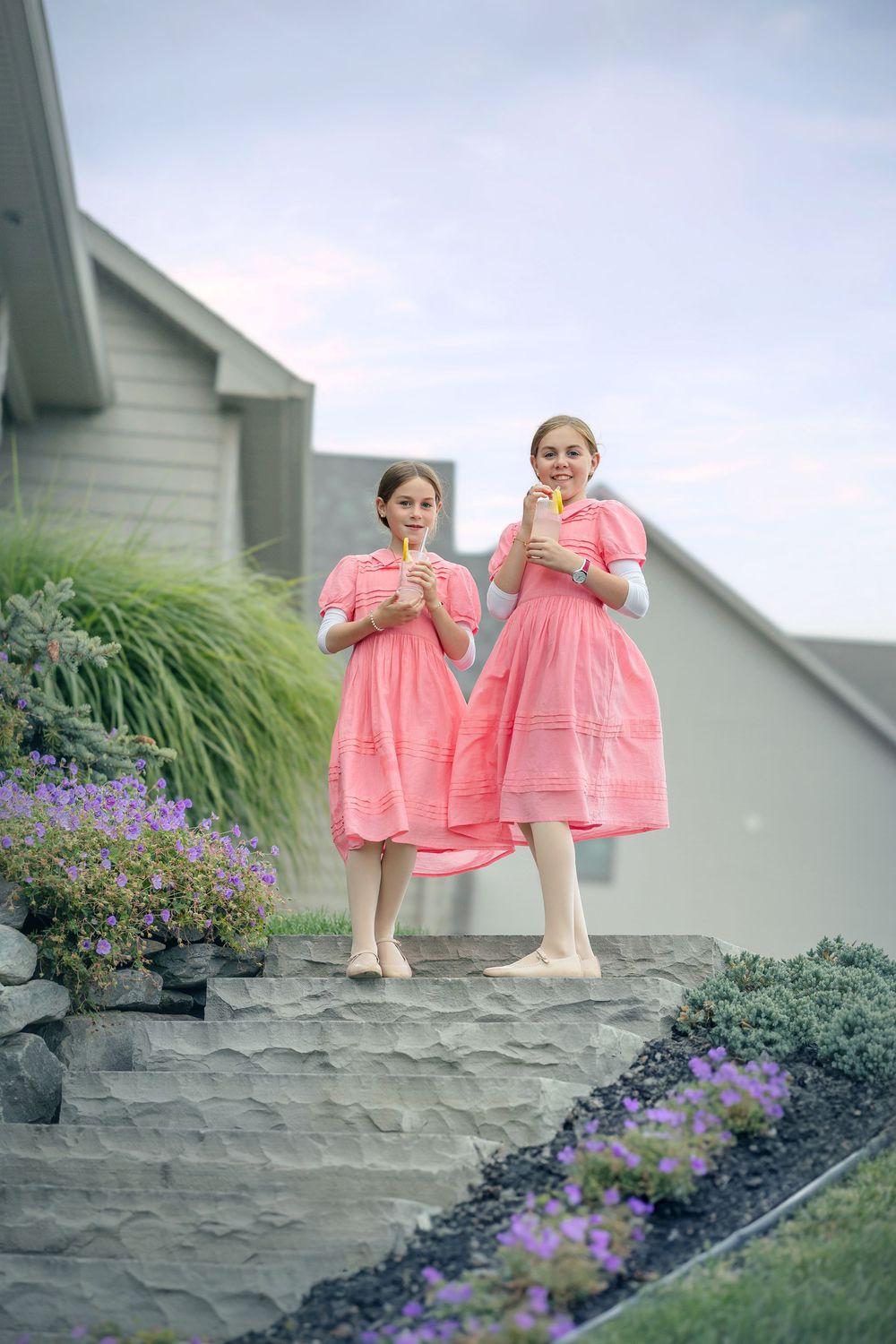Two figures in matching pink dresses stand on stone steps amid purple flowers and garden landscaping.