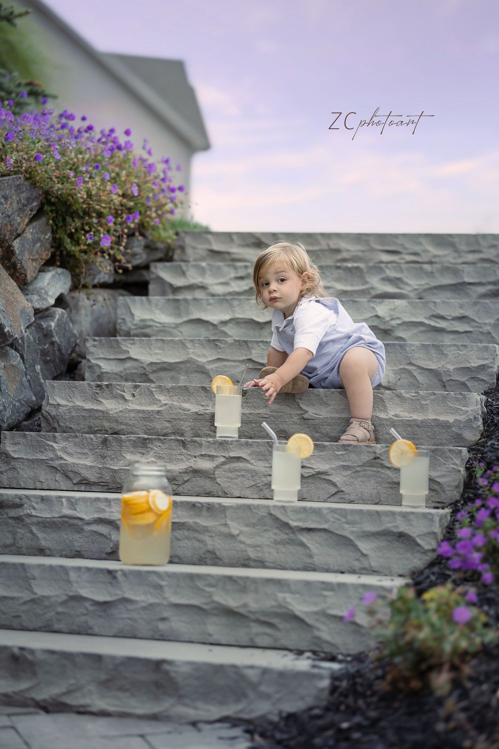 A toddler in a light-colored romper reaches for toys while playing on outdoor stone steps next to purple flowers.