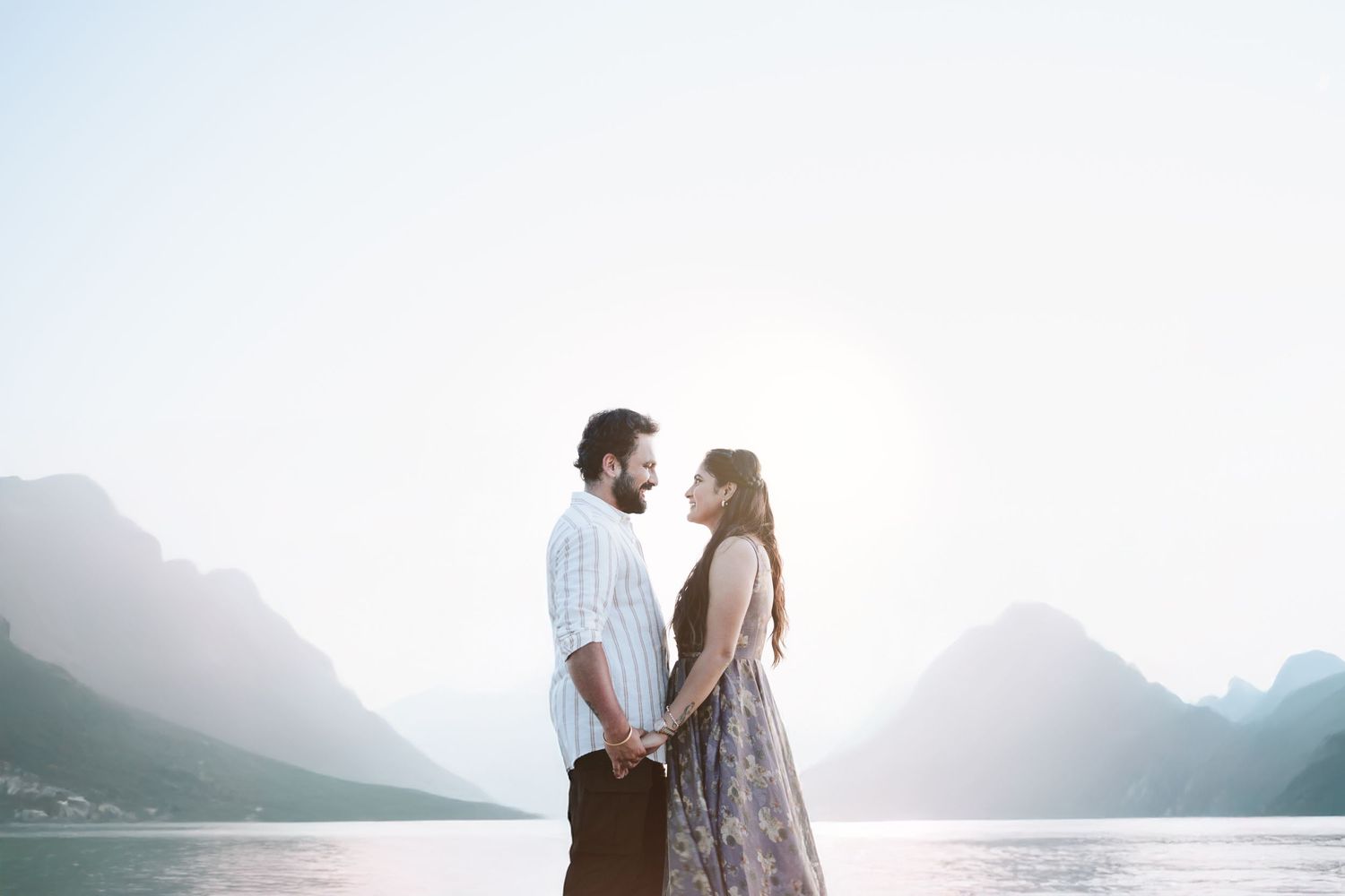 A couple stands together by a serene mountain lake at dusk with misty peaks in the background captured by Dallas Indian Wedding Photographer