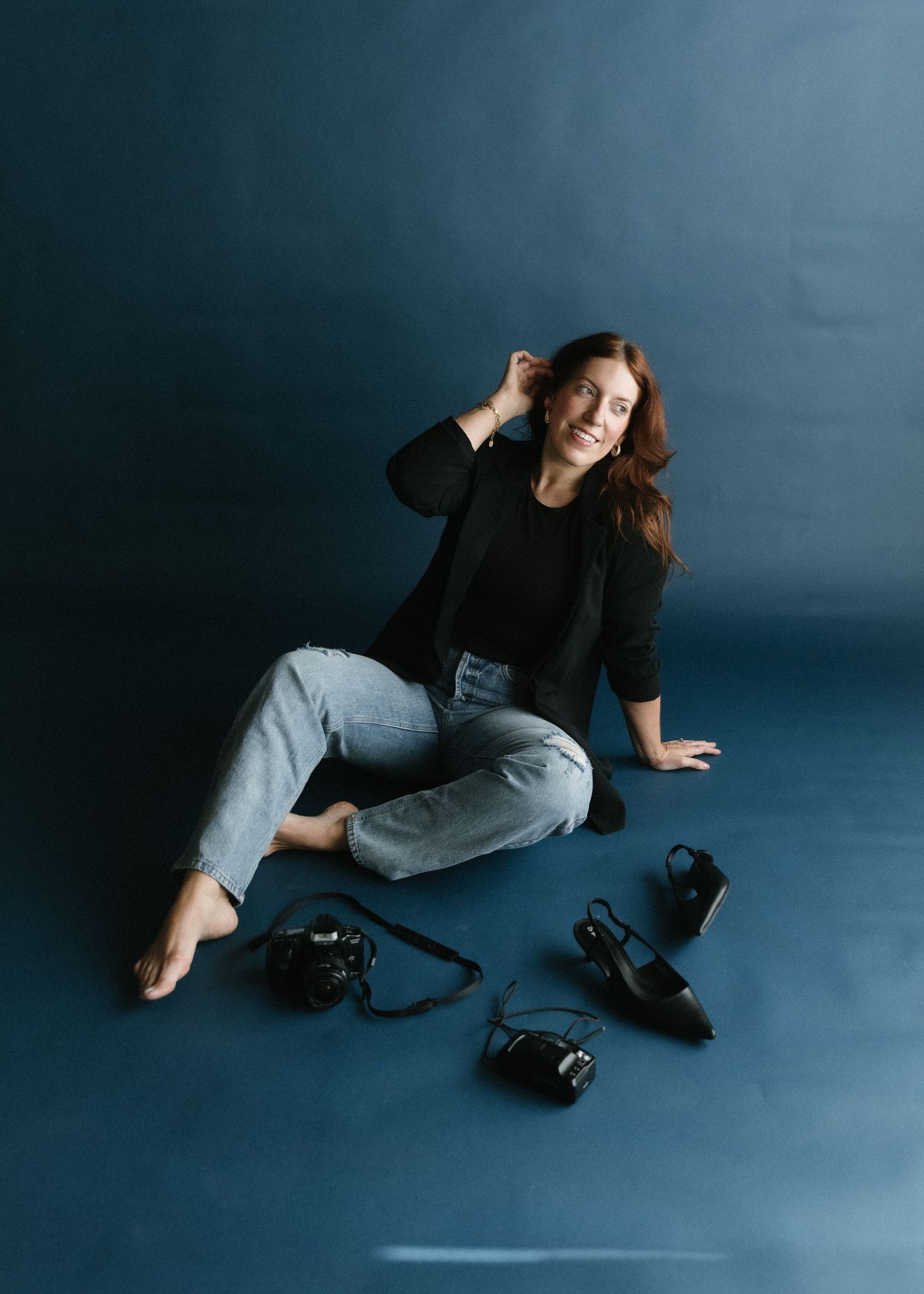 Photographer sitting on floor in casual black top and jeans with multiple cameras displayed around them on dark background.