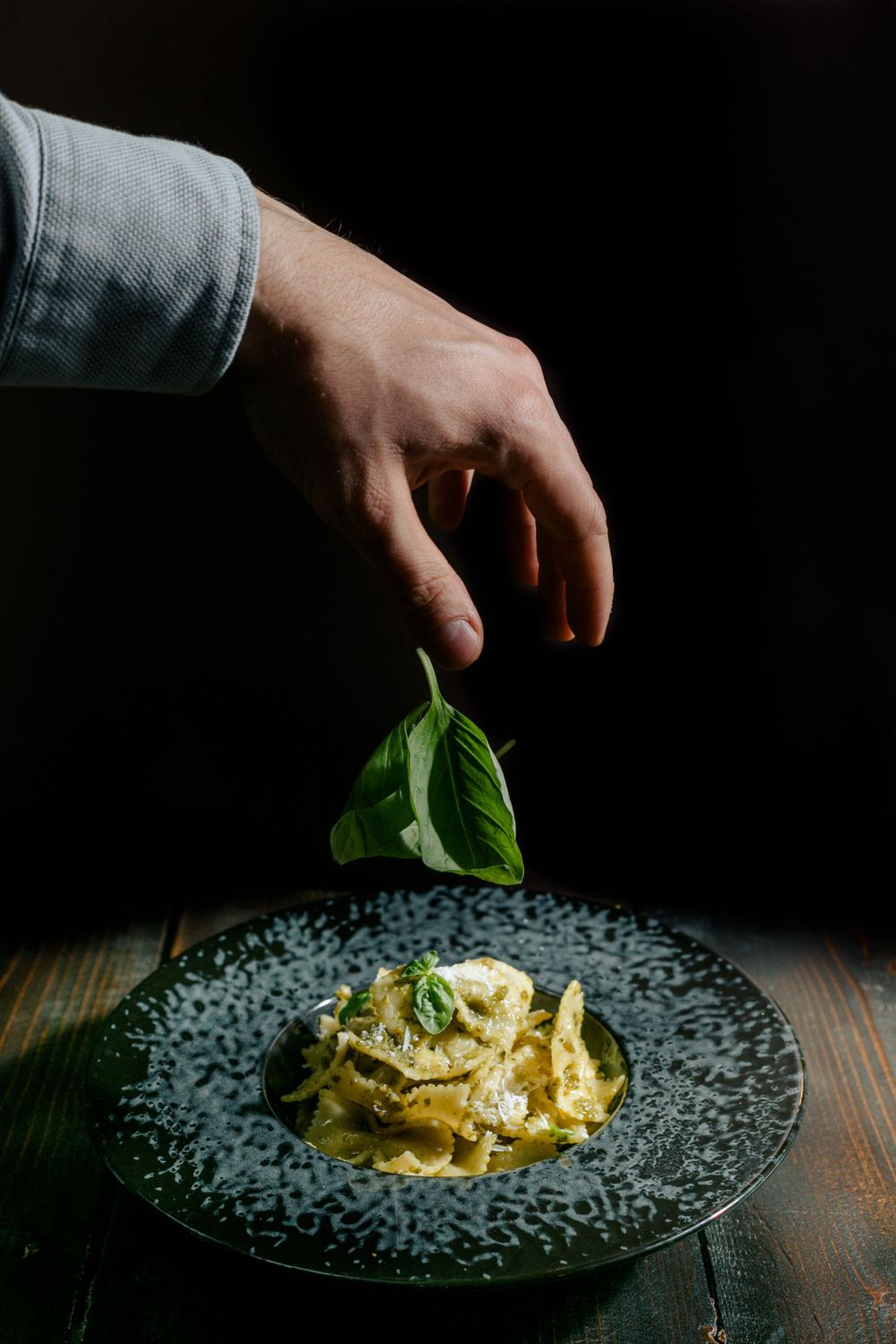 Garnishing creamy pasta dish with fresh basil leaf on textured black plate.