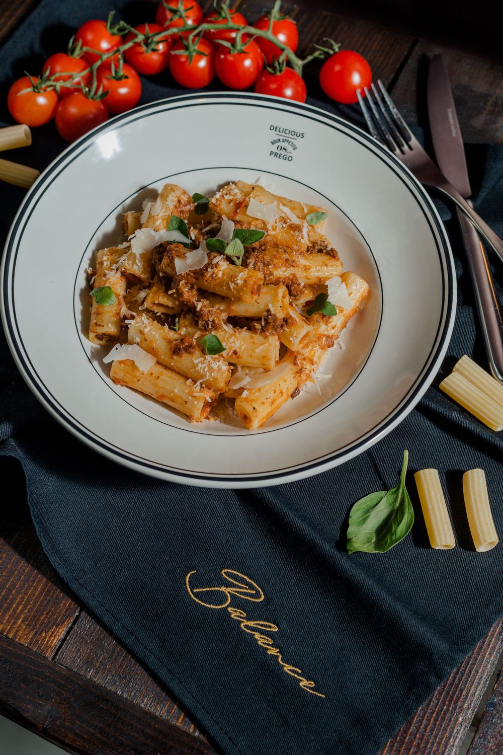 Creamy rigatoni pasta with herbs and tomatoes on white plate with dark napkin.