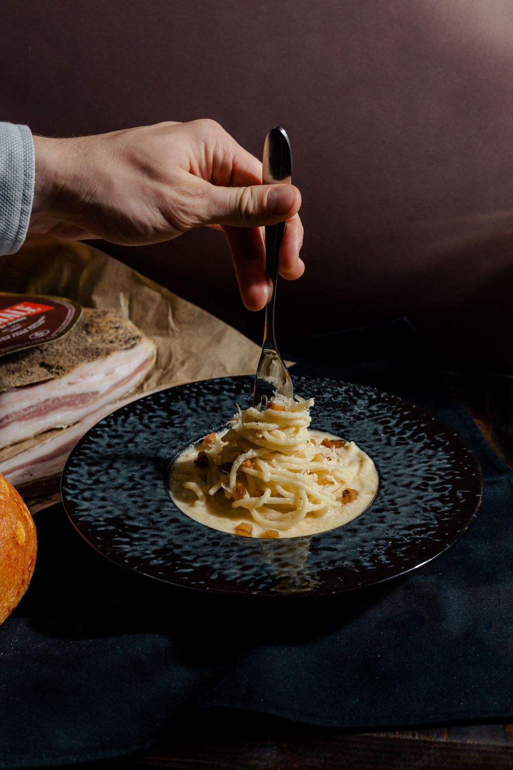 Twirling creamy carbonara pasta on black plate with fork in moody lighting.
