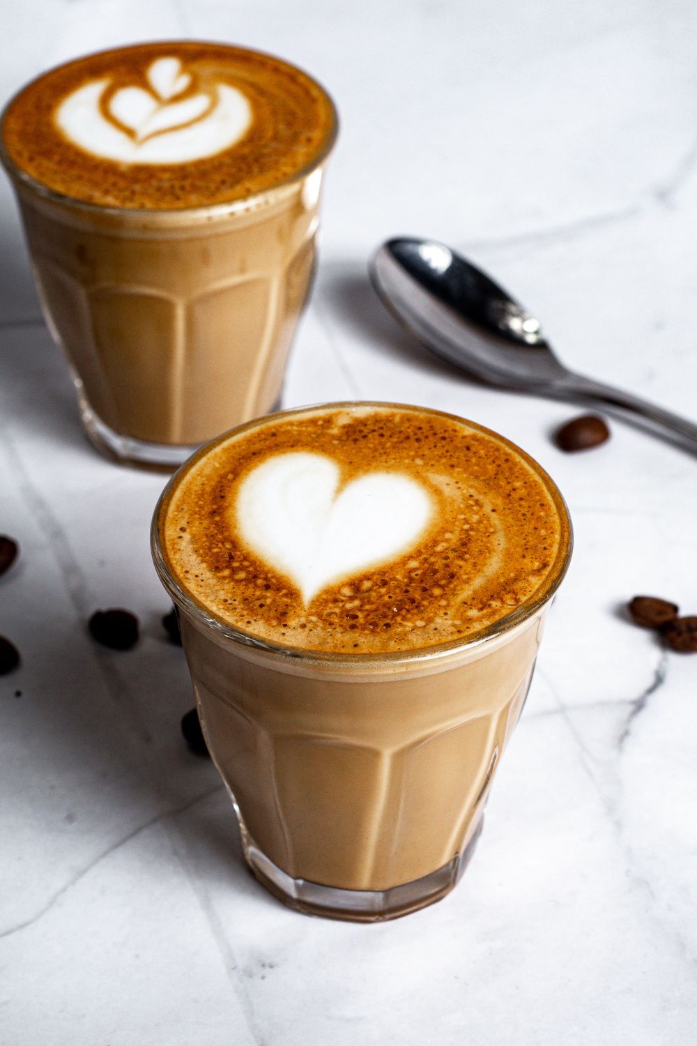 Two glasses of creamy latte with heart-shaped foam art on white marble surface next to coffee beans.