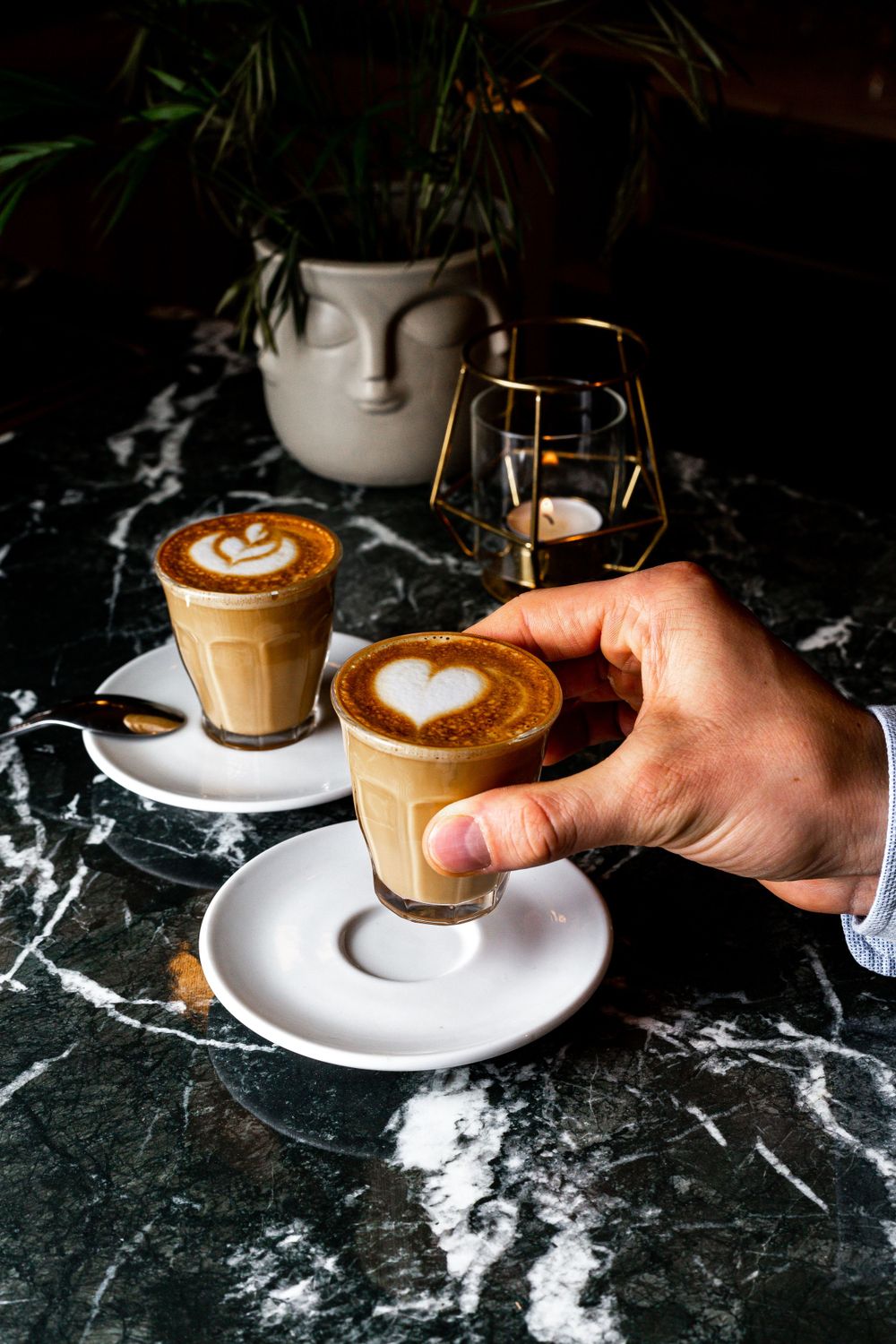 Two latte glasses with heart-shaped latte art on marble surface next to a white vase with plants.