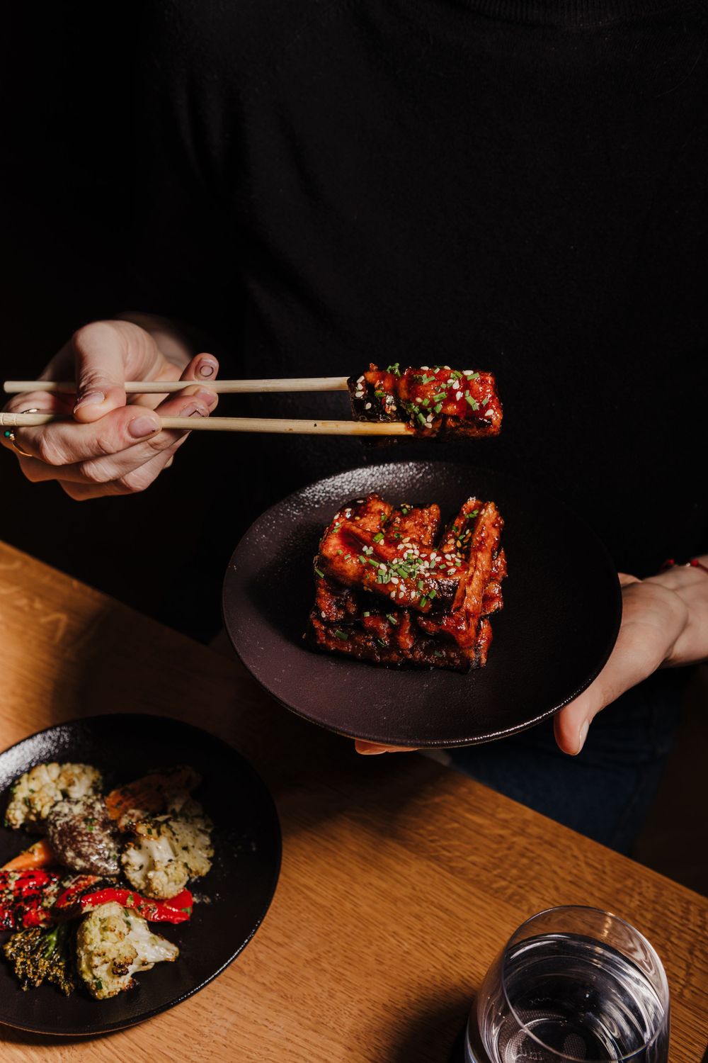Chopsticks lifting glazed Asian chicken from a dark ceramic plate with wine glass in view.