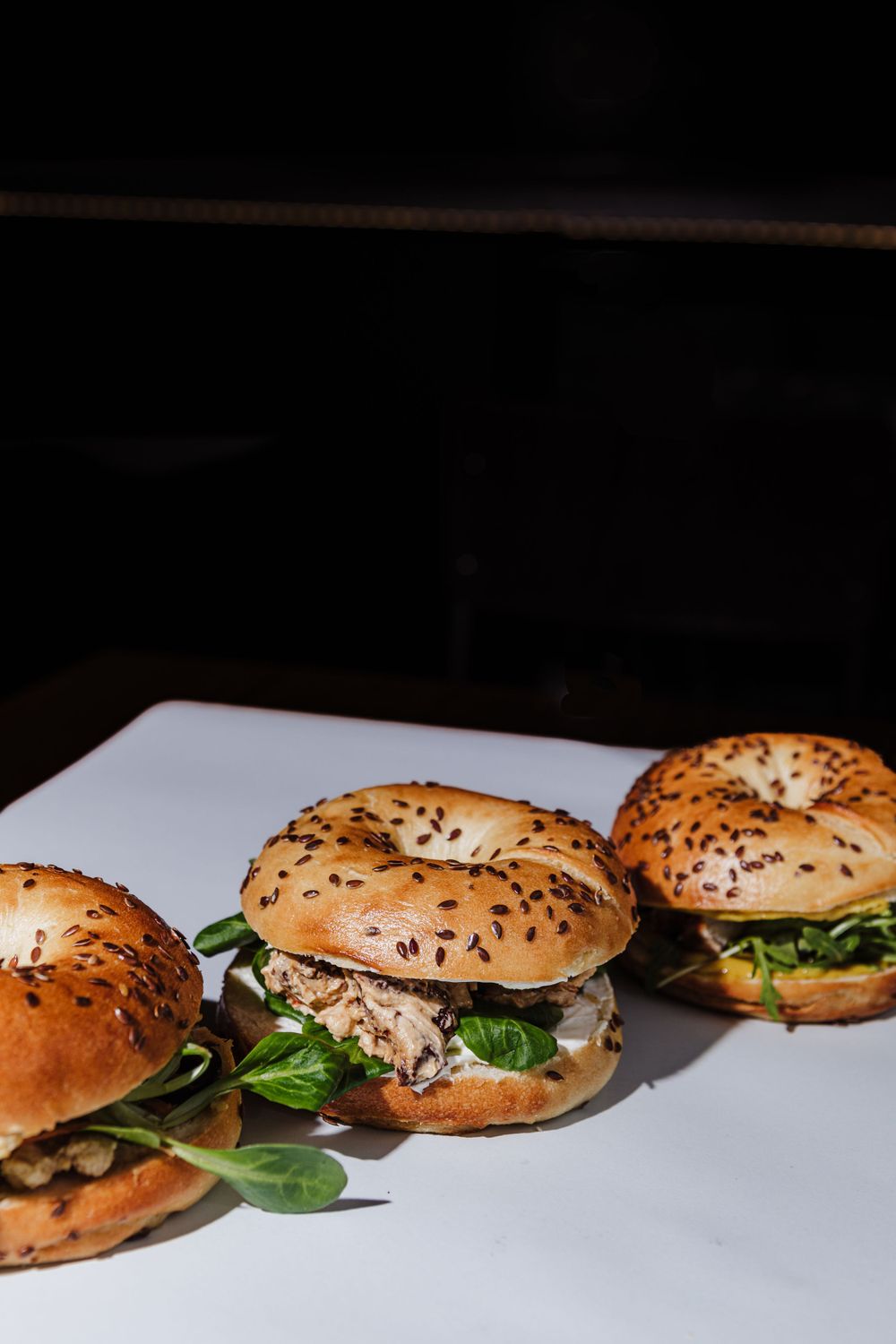 Three artisanal bagel sandwiches with fresh greens on white surface in dark setting.