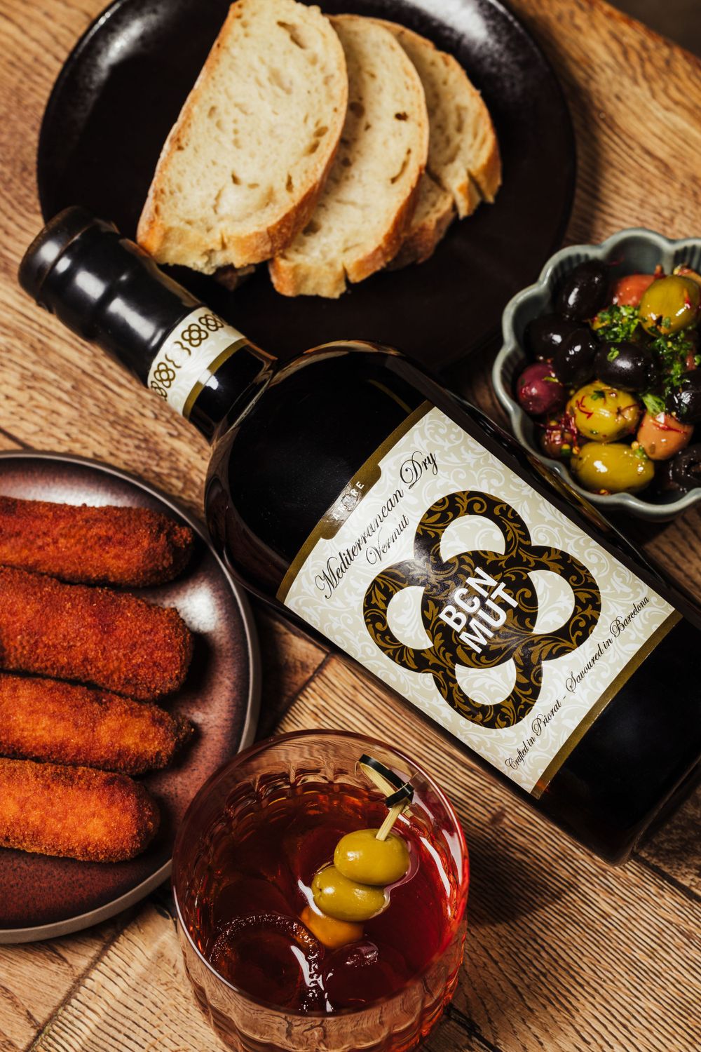Cocktail spread with sliced bread, croquettes and bottle of spirits on wooden table.