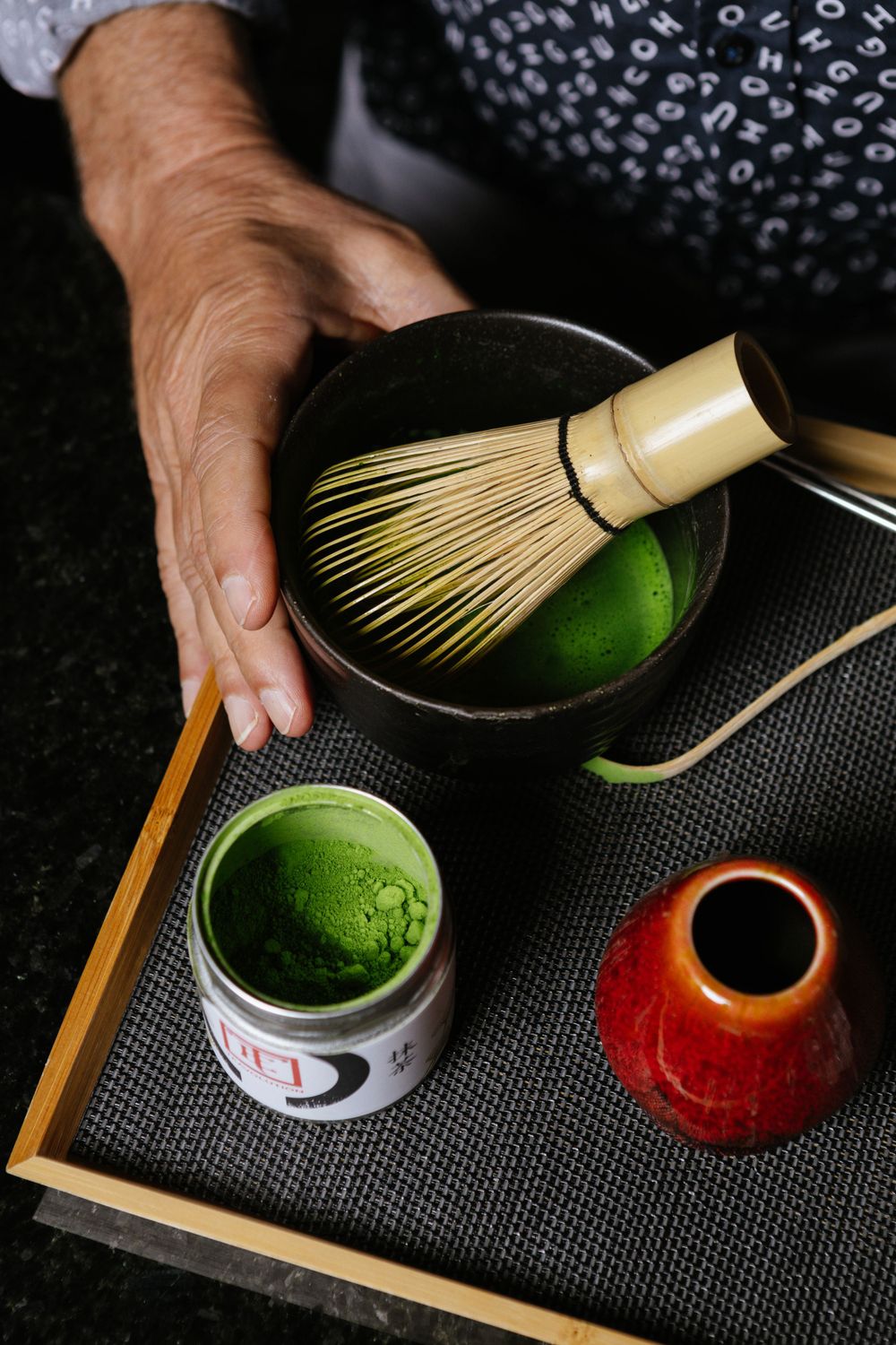 Traditional matcha tea preparation with bamboo whisk and green powder on dark tray.