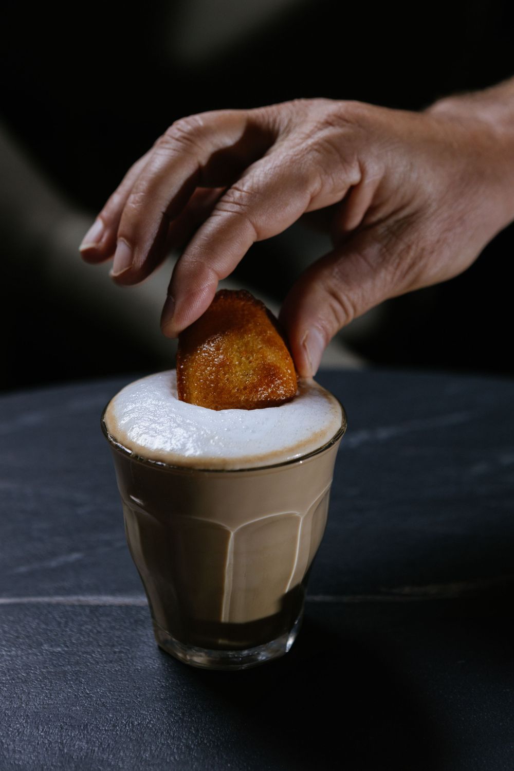 A cookie being dipped into a creamy latte drink with frothy milk on top.