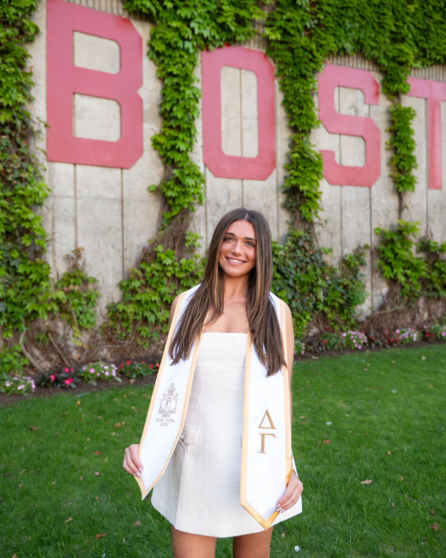 Graduate portrait in white dress standing in front of colorful flower bed and brick building.
