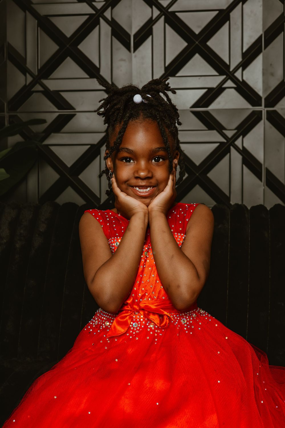 A young girl in a sparkling red ball gown smiles sweetly with hands on cheeks against a geometric backdrop.