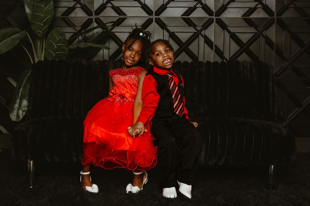 Two children dressed in matching red and black formal outfits pose together on a dark velvet sofa.
