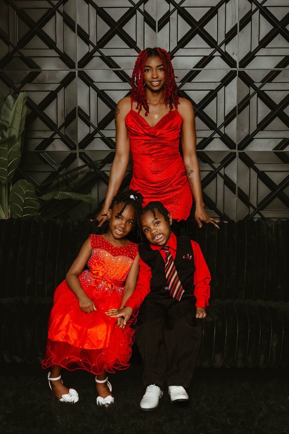 A woman in a red dress poses with two children dressed in red and black formal attire against a geometric backdrop.