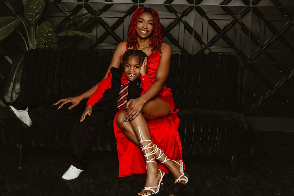 A woman in a red dress and child in black suit pose together on a dark velvet couch for a stylish photo.