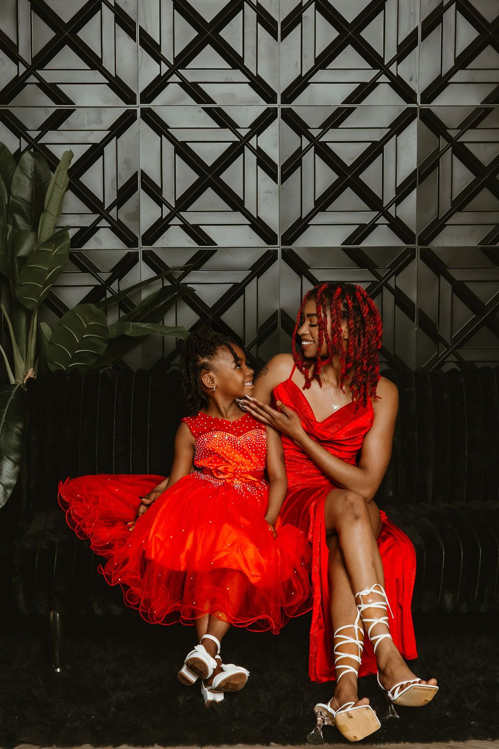 A woman and young girl in matching red dresses sit together smiling on a dark sofa against a geometric wall.