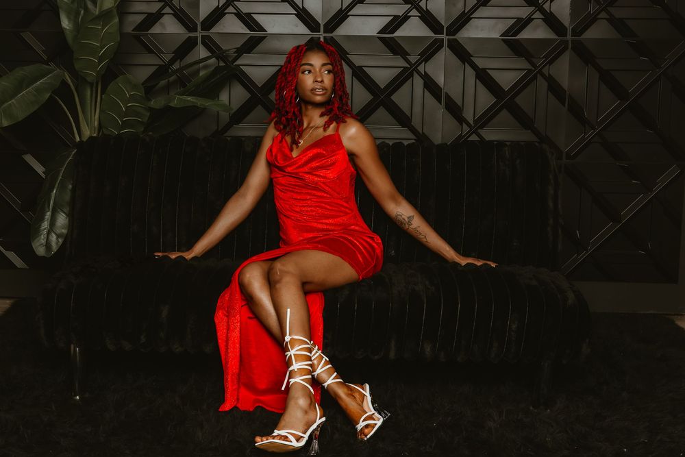 Woman in red satin dress and strappy heels poses confidently on dark velvet sofa in stylish interior.