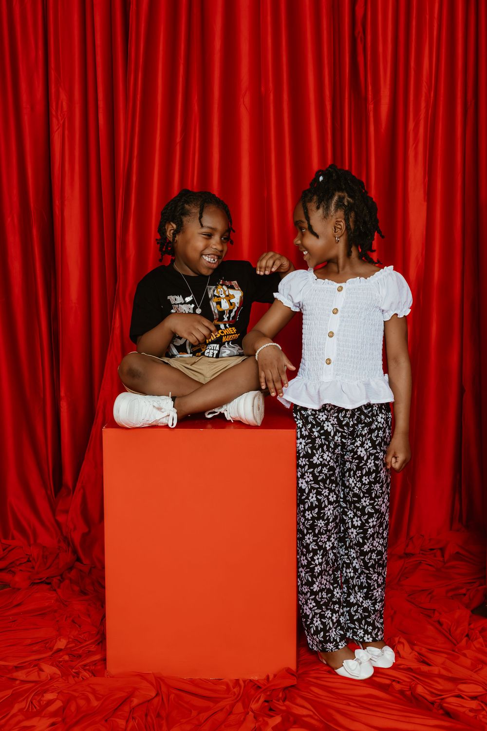 Two young girls laughing together in a studio, one sitting on a red cube, against a red curtain backdrop.