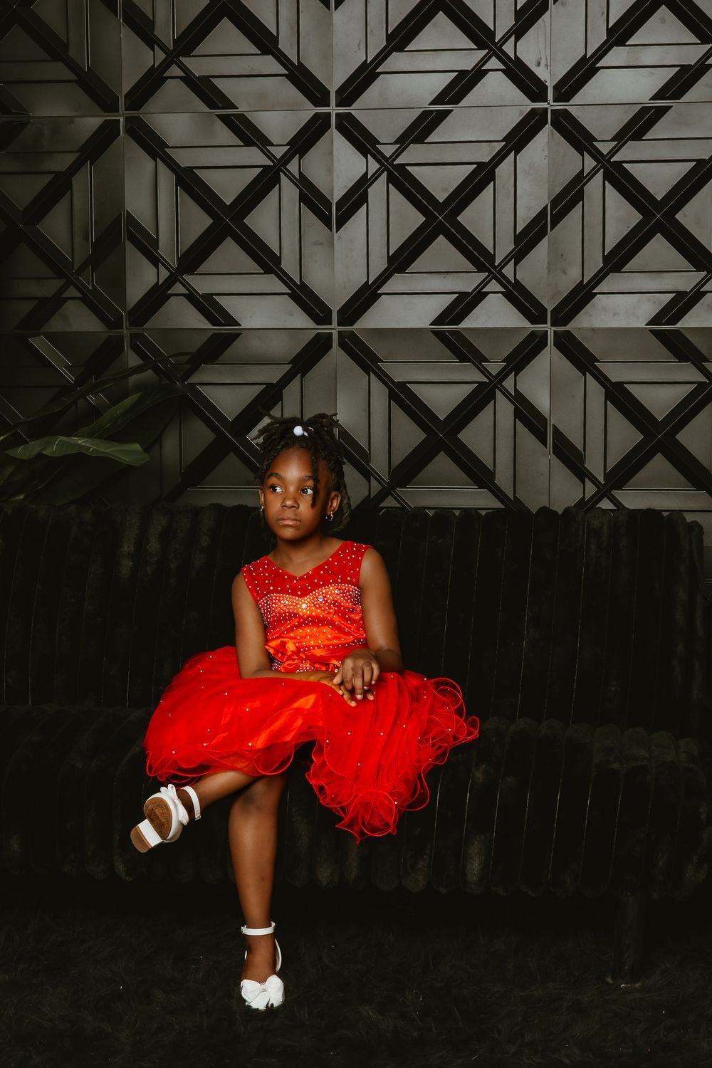 A young girl in a red tulle dress sits gracefully on a dark bench against a geometric patterned wall.