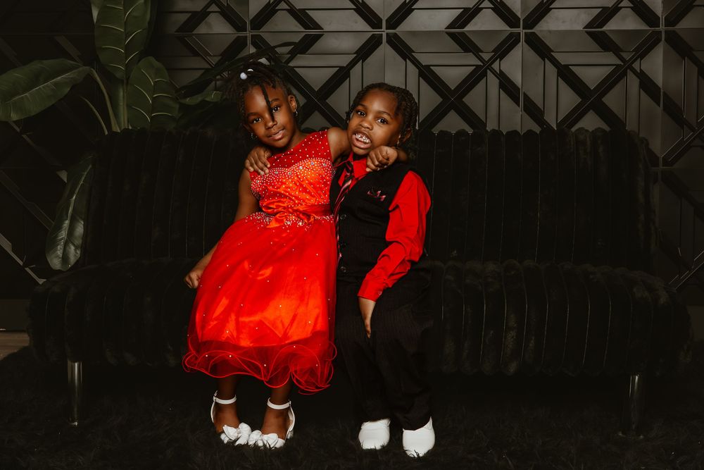 Two young children in red and black formal outfits pose together on a velvet sofa at an elegant event.