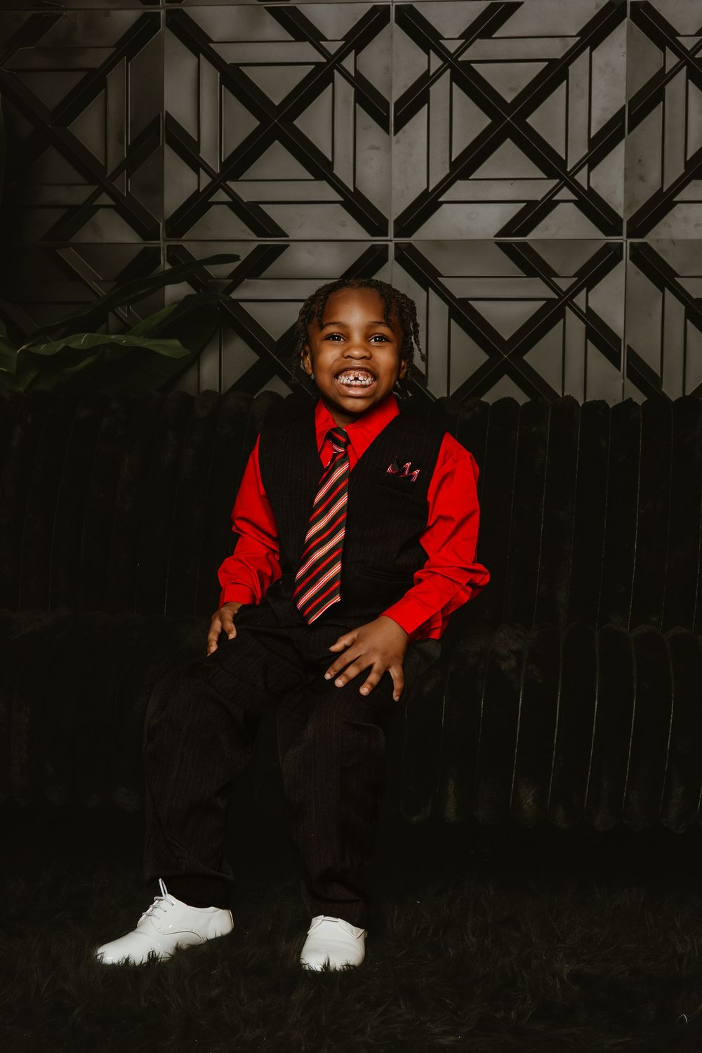 Young boy in red shirt, black vest and striped tie sits smiling for a formal portrait photo.