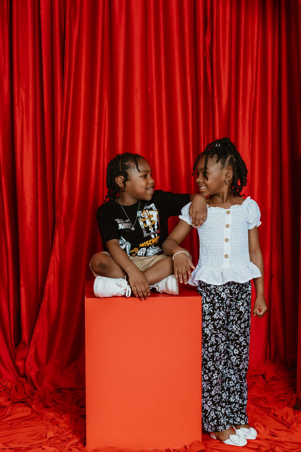 Two young children smiling at each other in front of a red curtain backdrop, sitting on an orange cube.