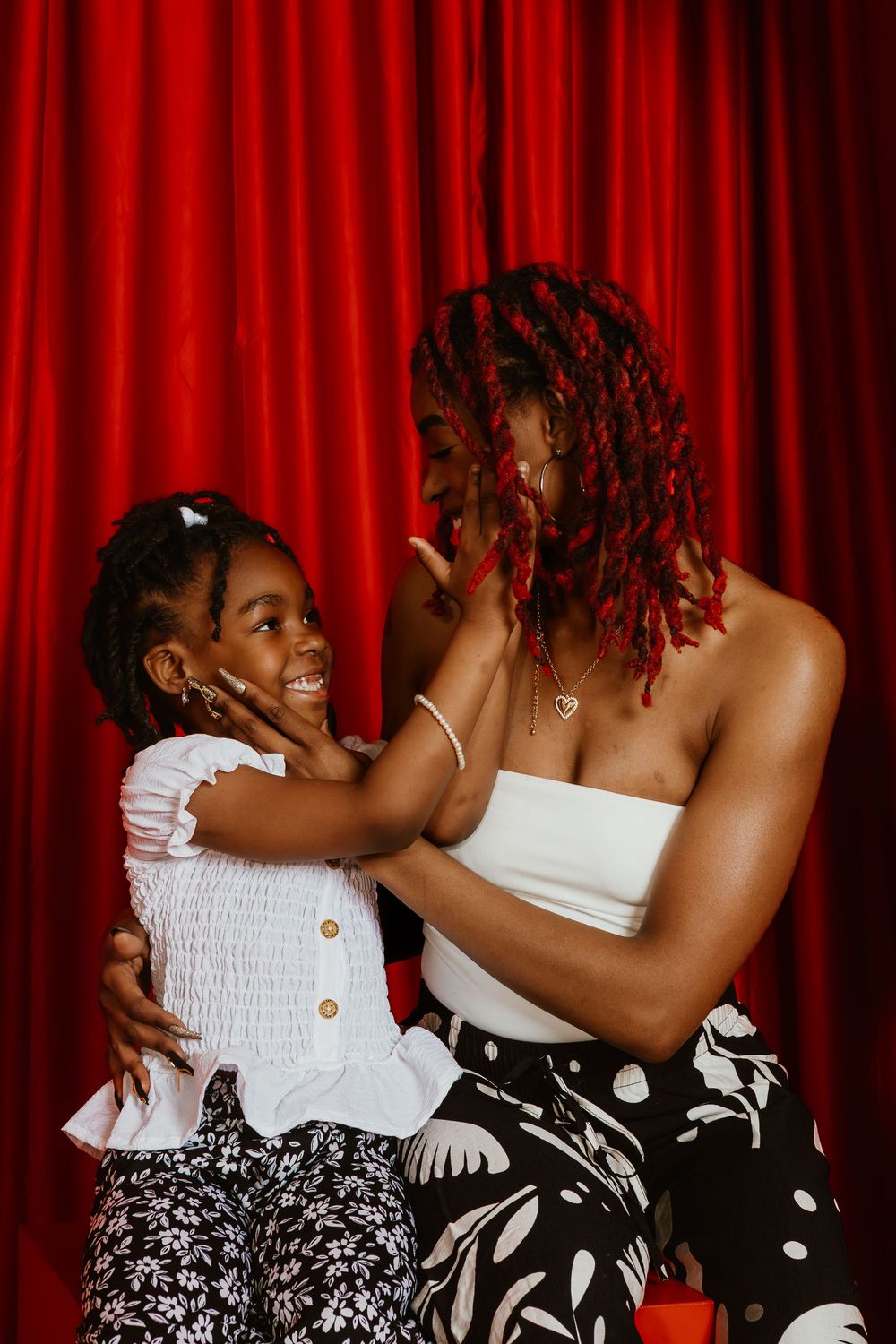 A joyful Black woman with red locs hugs a smiling little girl in white top, both in front of red curtain.